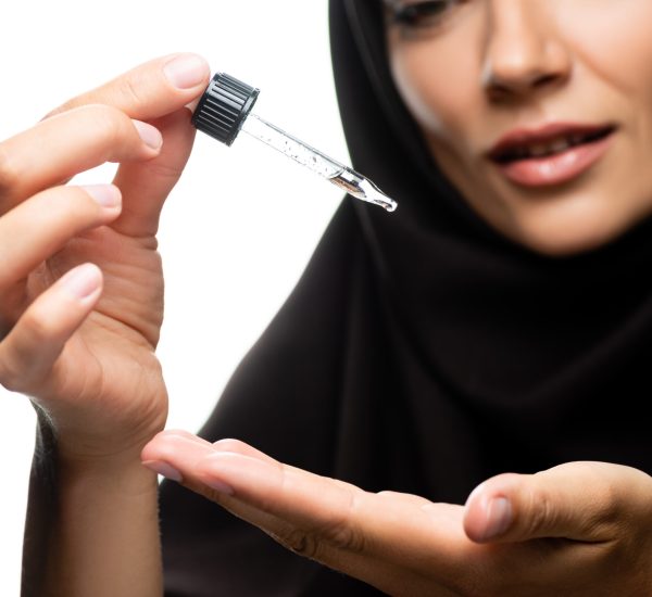 selective focus of young Muslim woman in hijab holding dropper with serum isolated on white